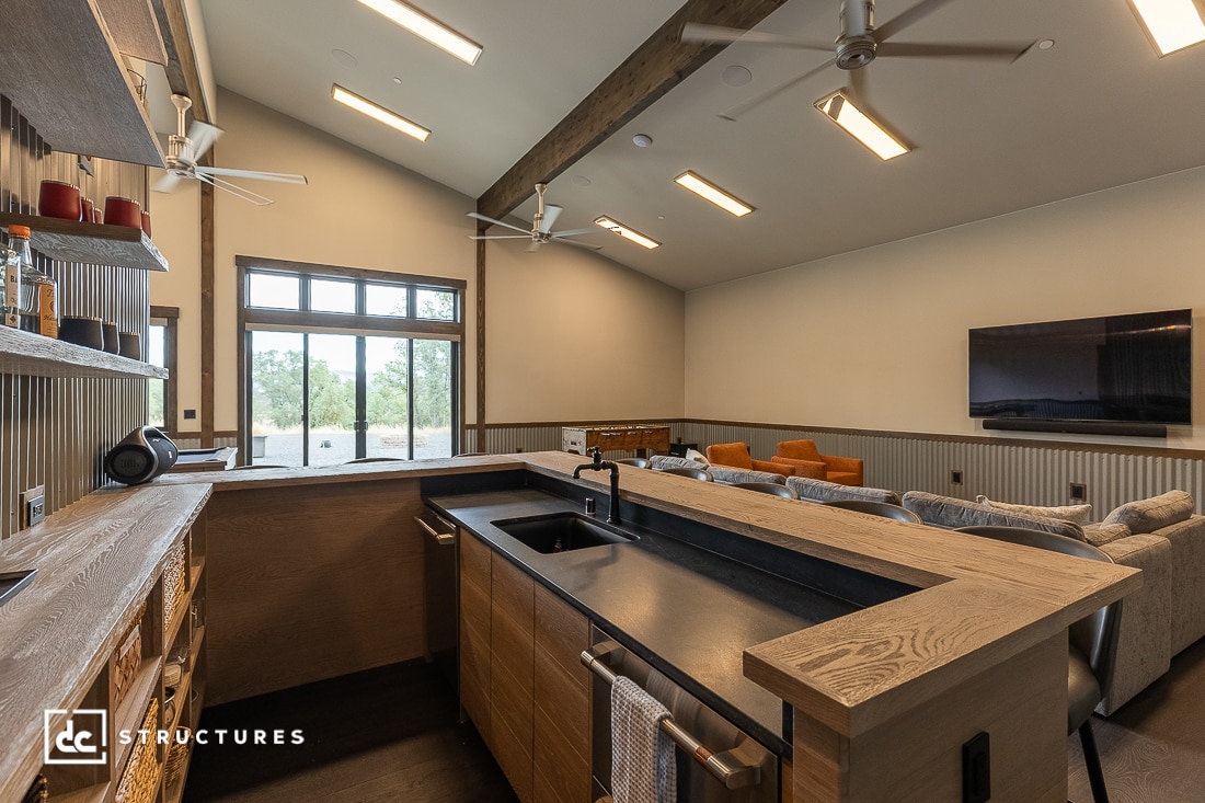 Modern living space with a wooden bar and sink in the foreground, a cozy seating area with couches and orange chairs, large windows letting in natural light, and a wall-mounted TV. Ceiling fans hang from a high, sloped ceiling.