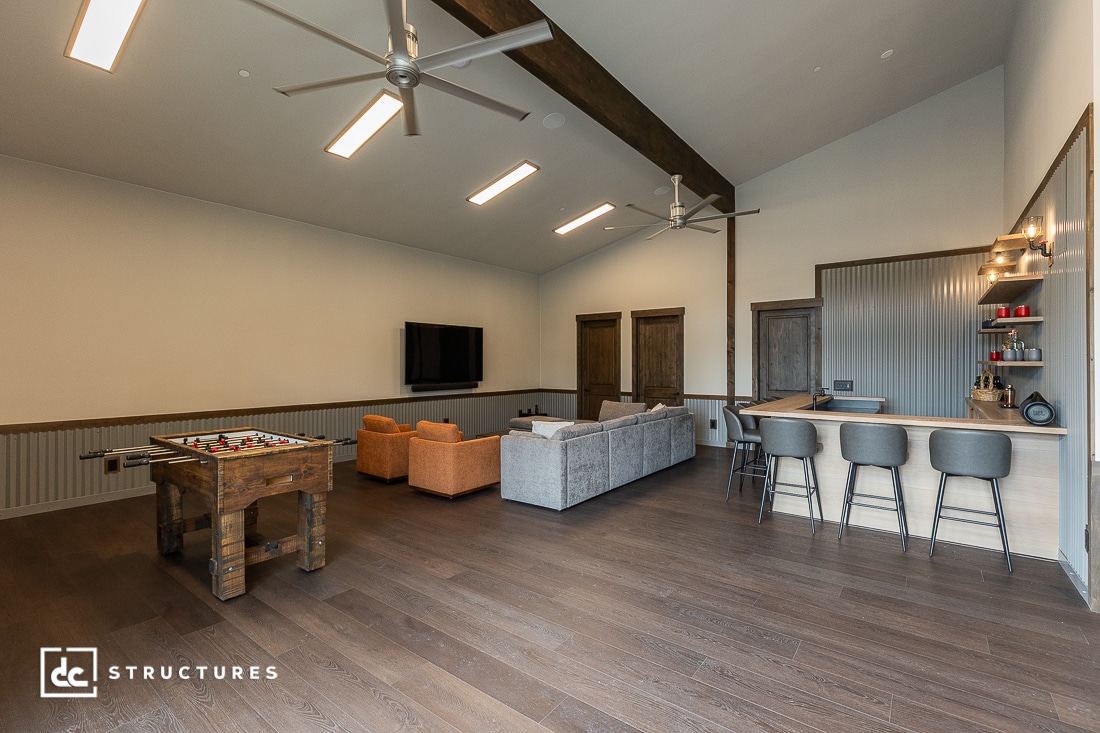 Spacious modern game room with wood floors, foosball table, bar area with stools, lounge seating, wall-mounted TV, and ceiling fans under a vaulted ceiling.