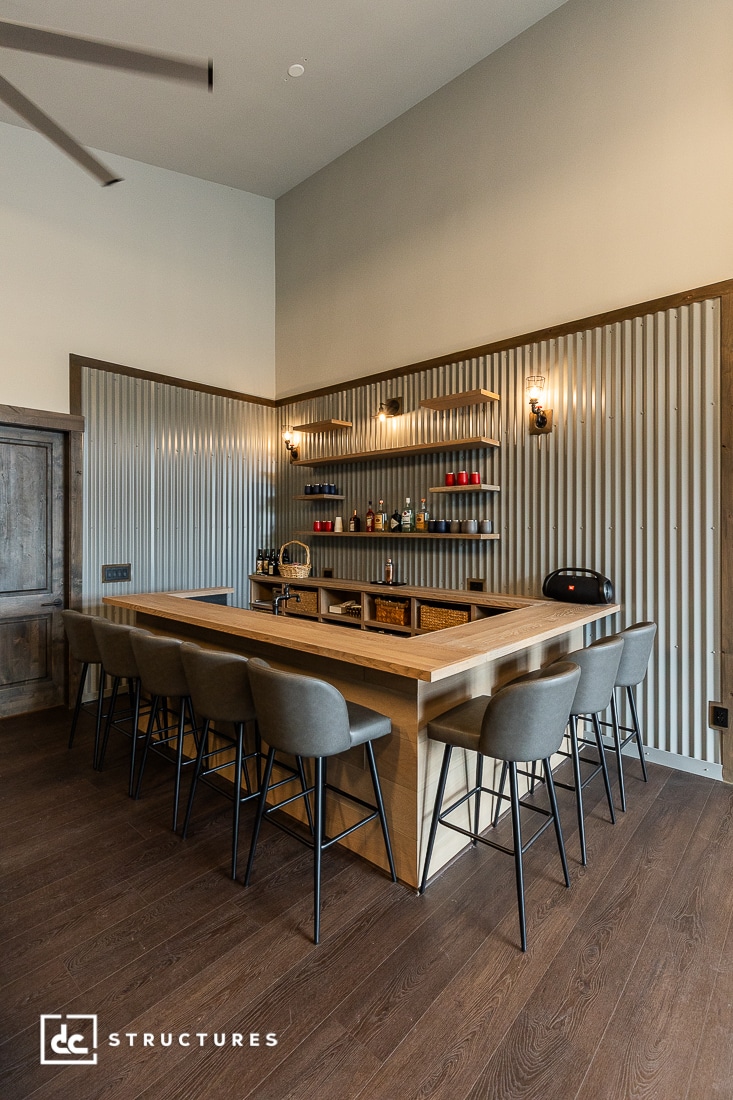 Modern home bar with a large wooden counter, gray upholstered barstools, and corrugated metal wall accents. Open shelves with drinks and décor, wood flooring, and wall-mounted sconces create a cozy, stylish atmosphere.