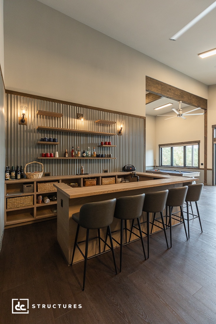 Modern home bar area with a wooden counter, four bar stools, open shelves displaying bottles and decor, and wicker baskets below. The space features corrugated metal wall panels and large windows.