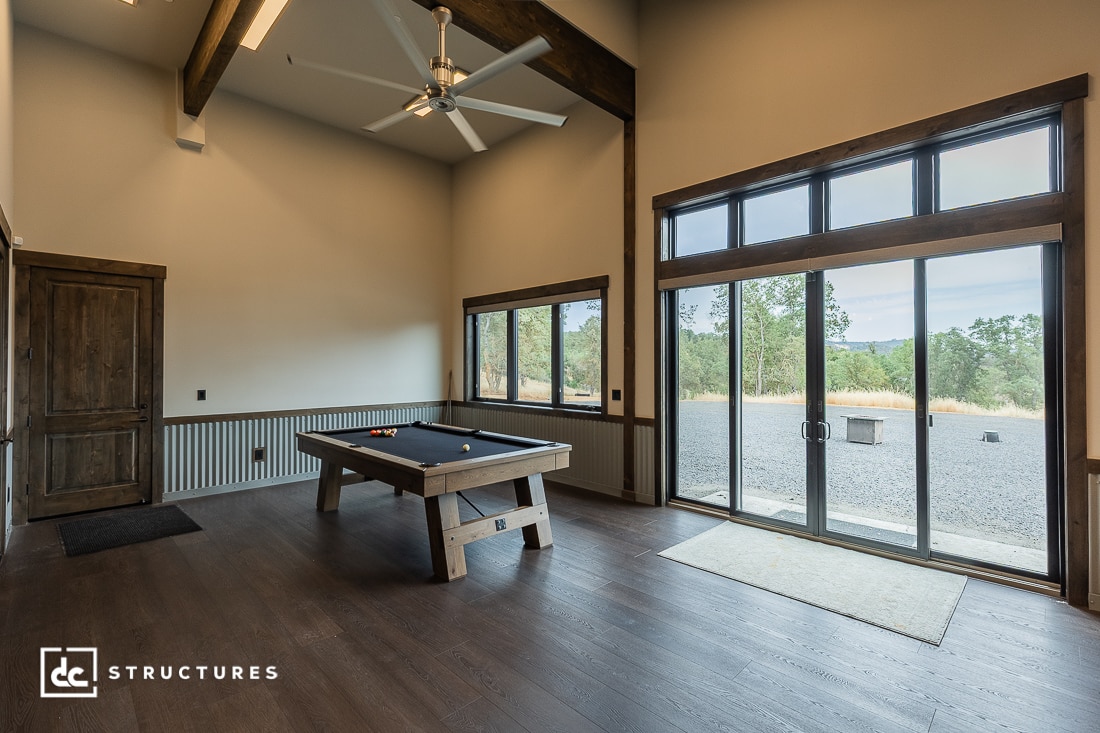 A spacious game room with a pool table, high ceiling with a fan, large windows and sliding glass doors overlooking a gravel patio and trees outside. The room features wood floors and rustic-style decor.