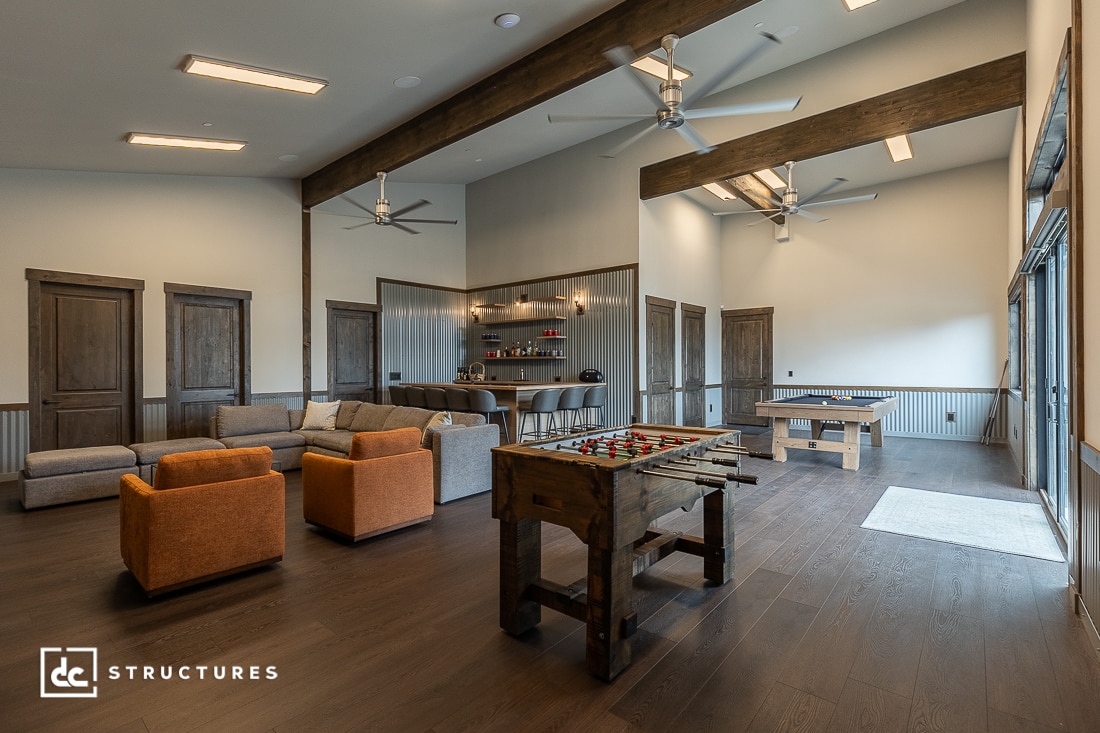 A spacious game room with large windows, featuring a foosball table, shuffleboard, sectional sofas, orange chairs, and a bar area with shelves and stools. The room has wood accents and ceiling fans.