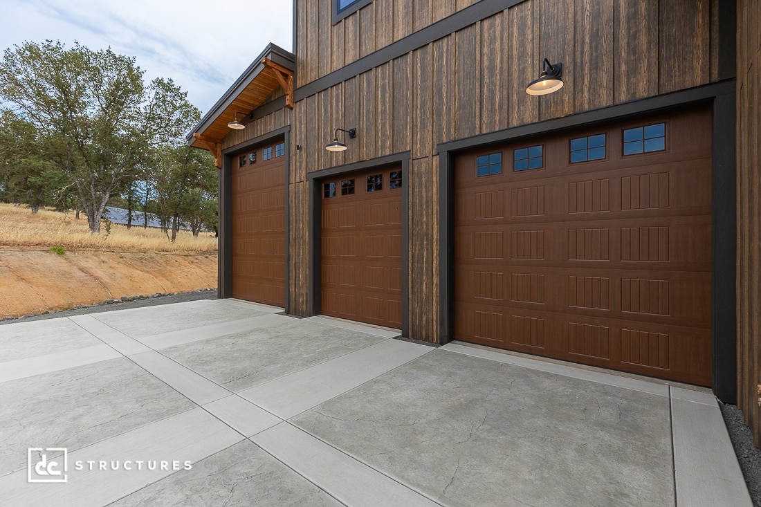 Three brown garage doors with small windows at the top, set in a modern wooden building with outdoor lights above each door. Concrete driveway and dry grassy landscape with trees in the background.
