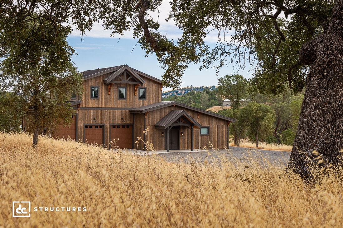 A modern two-story wooden house with three garage doors sits among tall dry grass and large oak trees, with rolling hills and trees visible in the background.