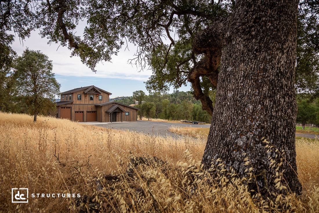 A large oak tree stands in the foreground of a dry grassy field, with a rustic two-story barn-style building in the background, surrounded by scattered trees under a partly cloudy sky.