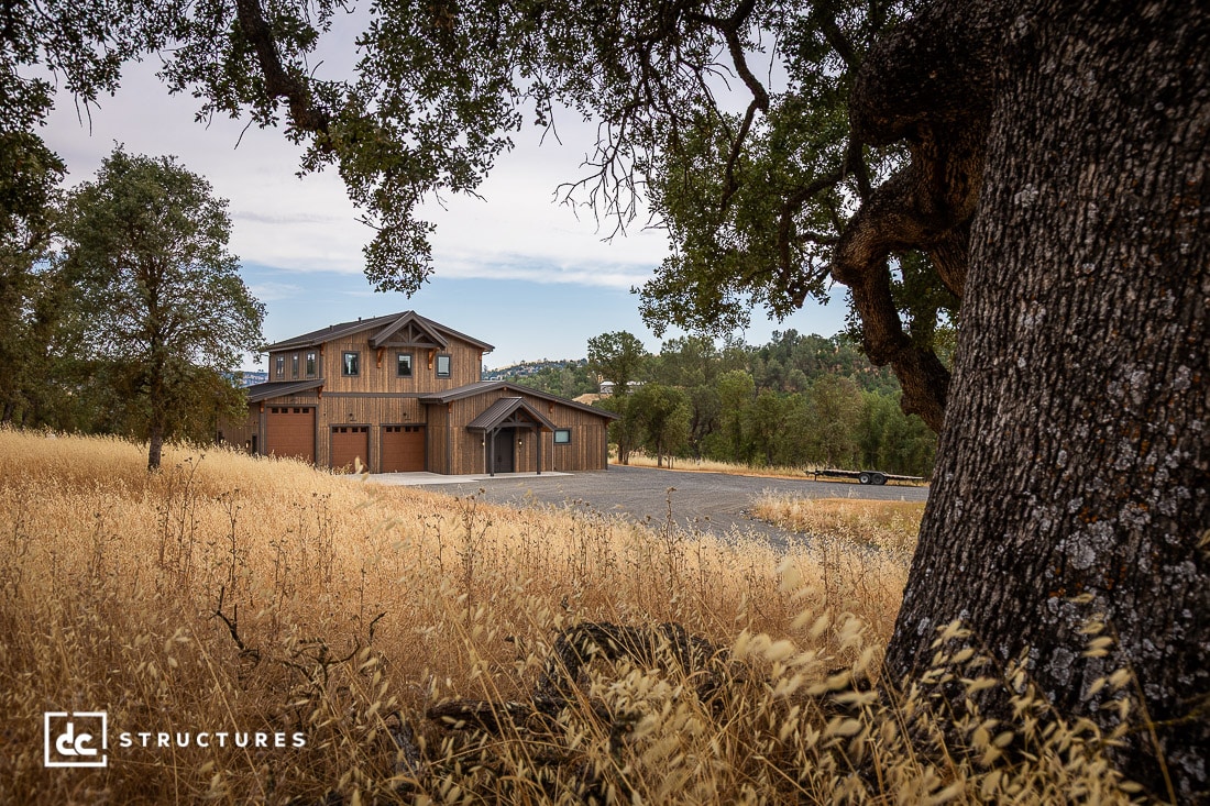A rustic two-story barn-style building with large garage doors sits amid golden dry grass and oak trees under a cloudy sky, with hills and scattered trees in the background.