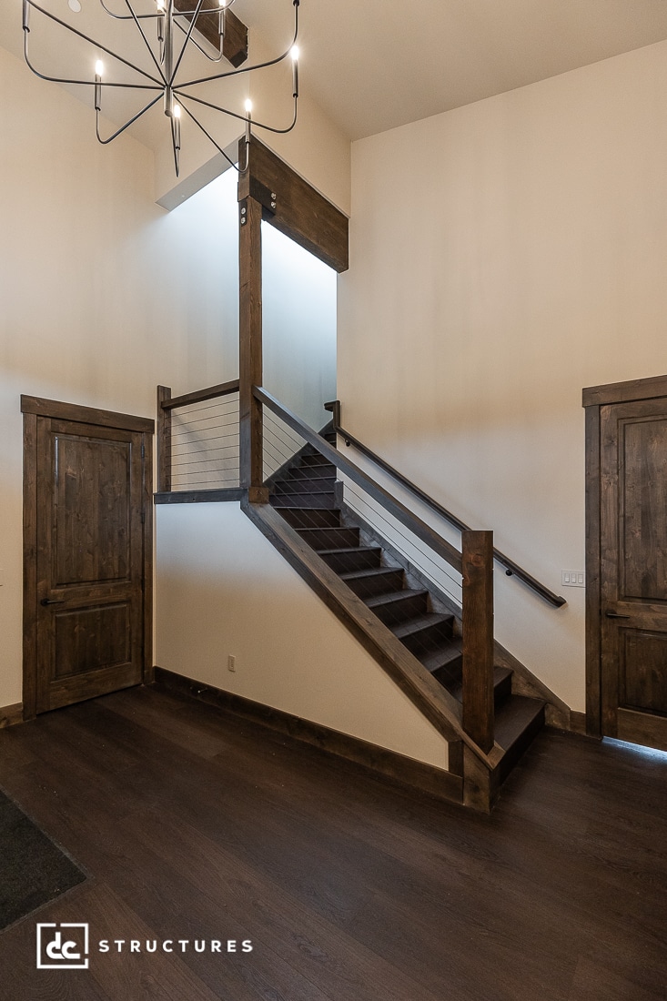 A modern staircase with dark wood steps and railings, set against white walls, leads to an upper level. Two wooden doors are on either side below, and a contemporary chandelier hangs above.
