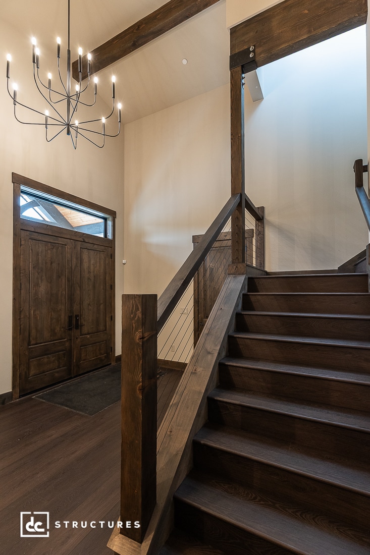 A modern entryway with dark wood double doors, a wooden staircase with cable railing, and a contemporary black chandelier; walls are light-colored, and the space feels open and airy.