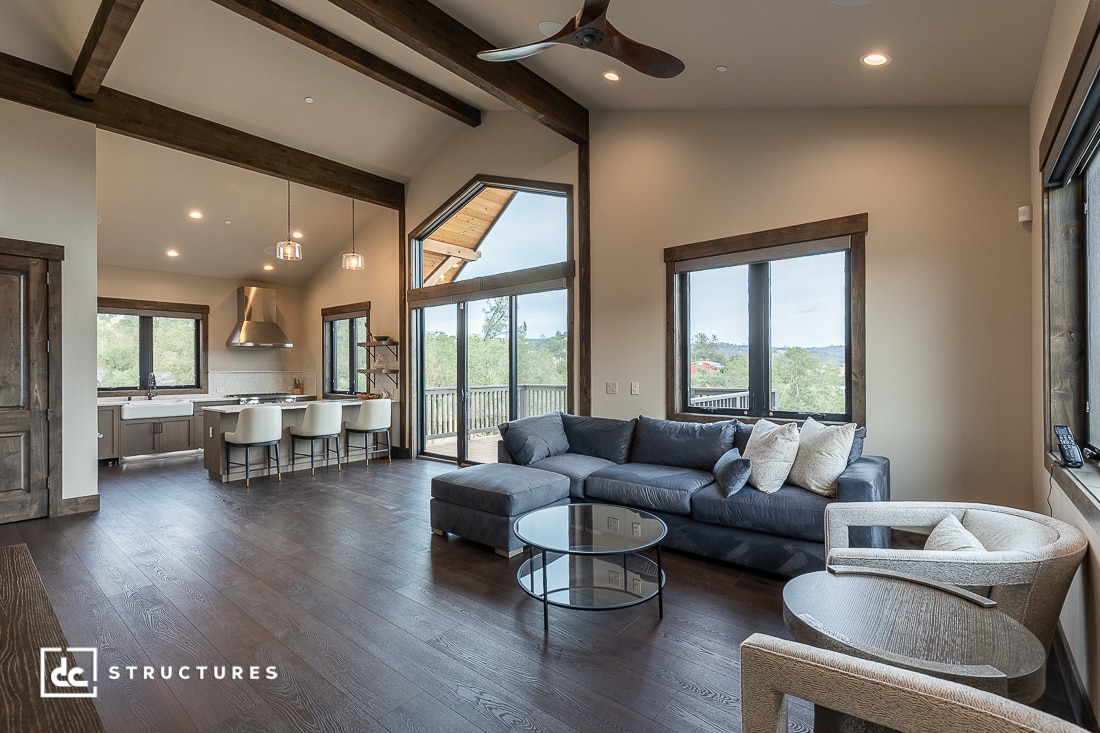 A modern open-concept living room and kitchen with dark wood floors, large windows, a gray sectional sofa, a round glass coffee table, and a dining area with white chairs and pendant lights.