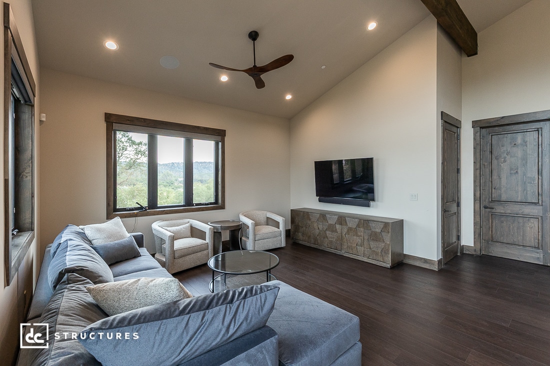 A modern living room with a gray sectional sofa, two beige armchairs, a wall-mounted TV, a geometric cabinet, dark wood floors, large windows with a nature view, and high vaulted ceilings with a ceiling fan.