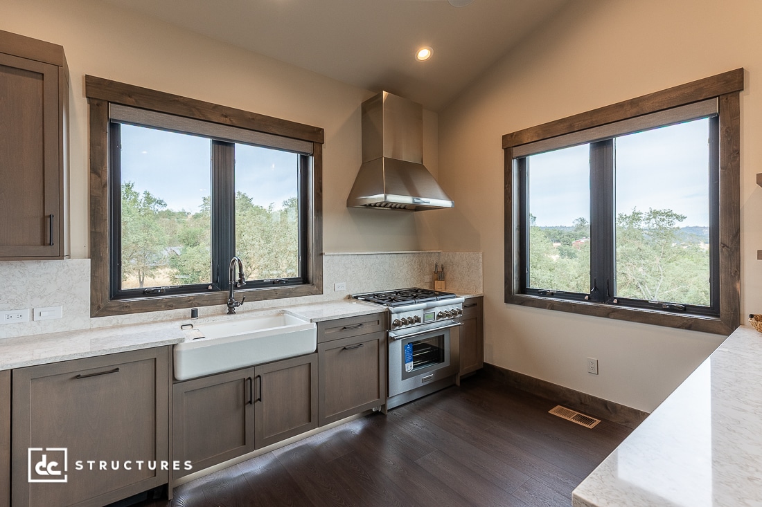 A modern kitchen with large windows, a farmhouse sink, stainless steel range and hood, wood cabinets, and marble countertops. Trees and greenery are visible outside the windows.
