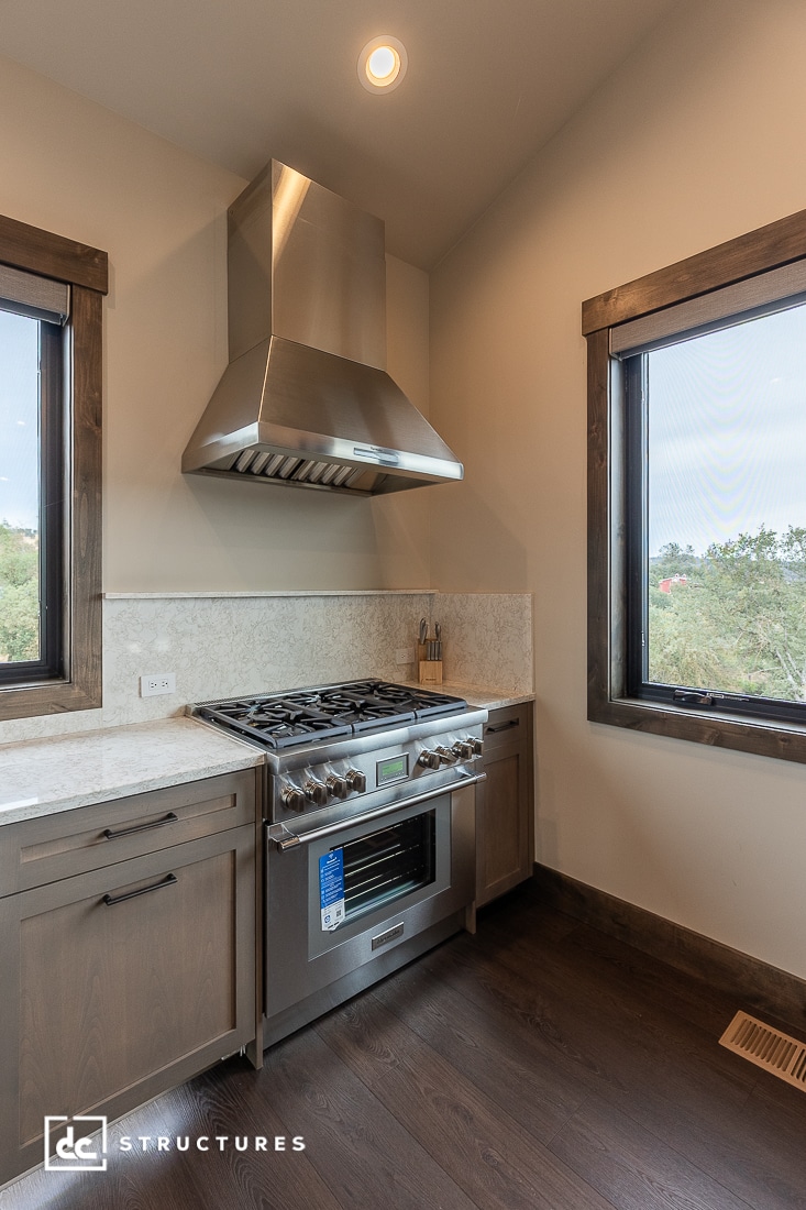 A modern kitchen features a stainless steel gas stove with a large vent hood, surrounded by brown cabinets, marble backsplash, and two large windows with dark wood frames letting in natural light.