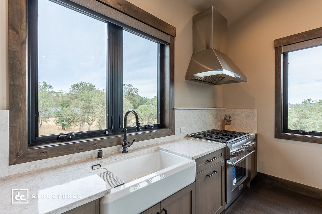Modern kitchen with a large farmhouse sink under a wide window, stainless steel stove and range hood, marble countertops, and wooden cabinets. Trees and a field are visible outside the windows.