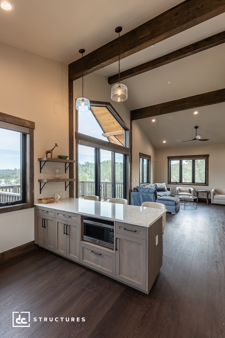Modern open-concept living area with large windows, exposed wooden beams, pendant lights, and a kitchen island featuring a built-in microwave. A seating area with sofas and a view of trees are visible in the background.