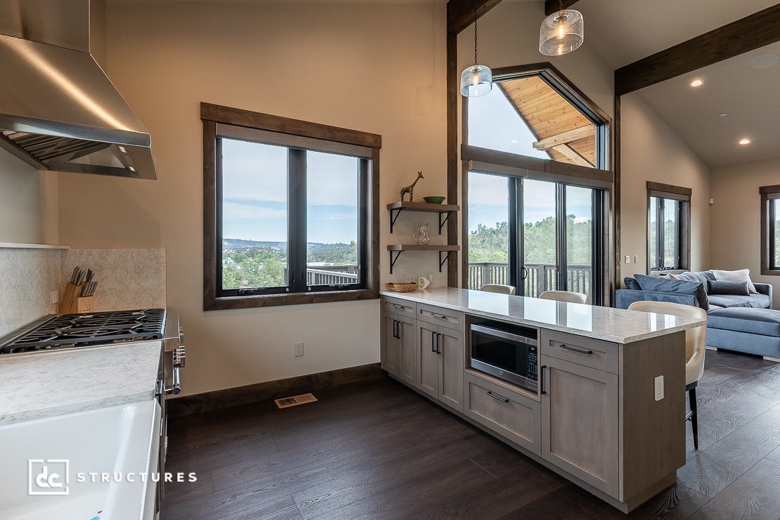 Modern kitchen with wood floors, large windows, an island with built-in microwave, and pendant lights. Open layout with a view into the living area and outdoor balcony, surrounded by greenery and sky.