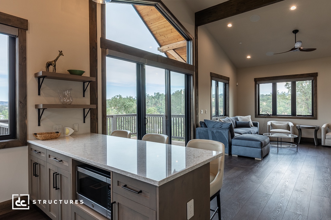 Modern open-concept kitchen and living room with large windows, a kitchen island with stools, light cabinetry, and a cozy seating area. Natural light fills the space, highlighting wooden and neutral tones.