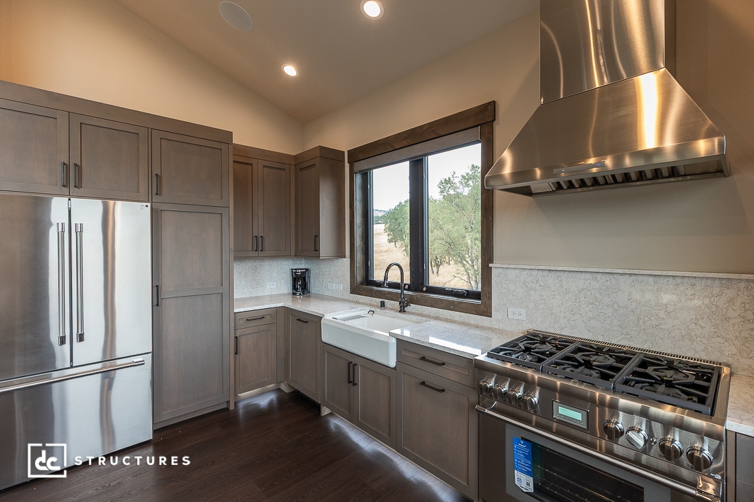 Modern kitchen with stainless steel appliances, a farmhouse sink under a large window, wood cabinets, dark flooring, and a tile backsplash.