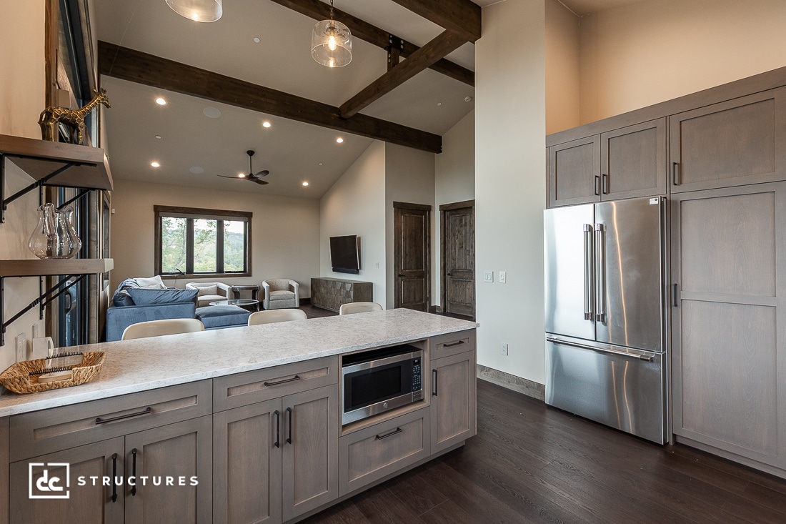 Modern open-concept kitchen and living room with wood beams, gray cabinets, stainless steel appliances, a kitchen island, and a cozy seating area with large windows and a wall-mounted TV.