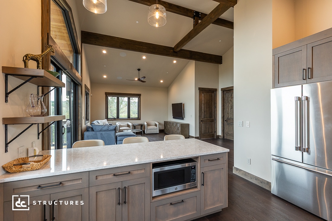 Modern open-concept kitchen and living room with exposed wood beams, light wood cabinetry, stainless steel appliances, and large windows overlooking a seating area with a TV and blue sofas.