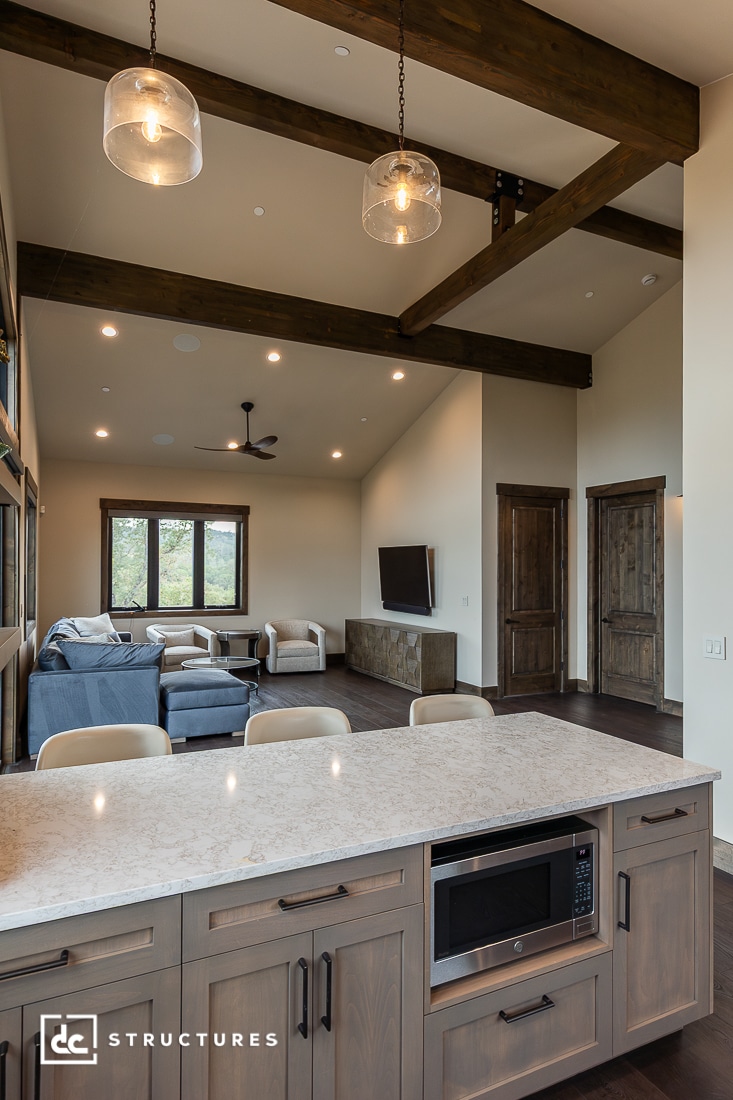 Spacious living area with exposed wooden beams, pendant lights, light wood cabinets, a marble countertop with built-in microwave, and a seating area with sofas, a TV, and large windows.