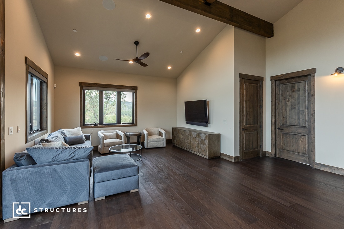 A spacious living room with dark wood floors, beige walls, a sectional sofa, two armchairs, a wall-mounted TV, and large windows. The room features wood accents and modern ceiling lights.