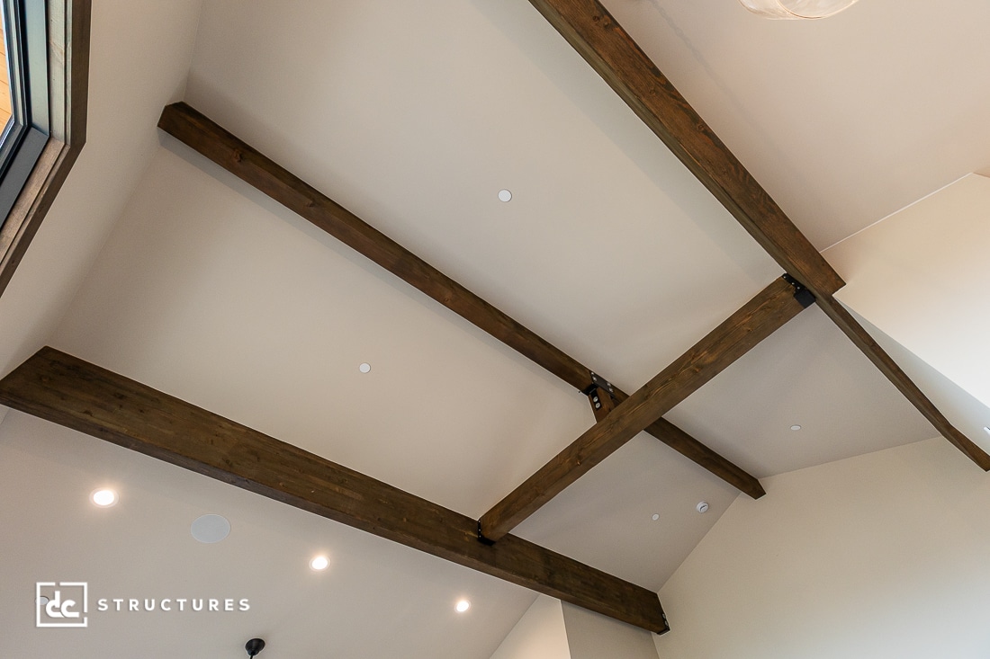 Exposed wooden beams cross a high, cream-colored ceiling with recessed lighting. The photo is taken from below, showing the beams’ rustic texture and simplicity. A window frame and part of a pendant light are visible.