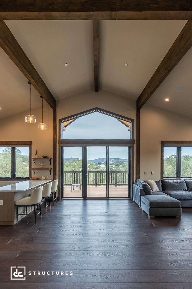 Open living space with dark wood floors, exposed beams, and large glass doors leading to a deck with scenic outdoor views. Modern furniture includes a gray sectional sofa and white barstools along a kitchen island.
