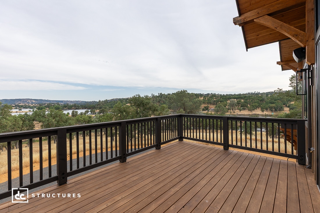 Spacious wooden deck with black railing overlooks a scenic landscape of rolling hills, trees, and a distant body of water, under a cloudy sky. Part of a wooden structure is visible on the right.