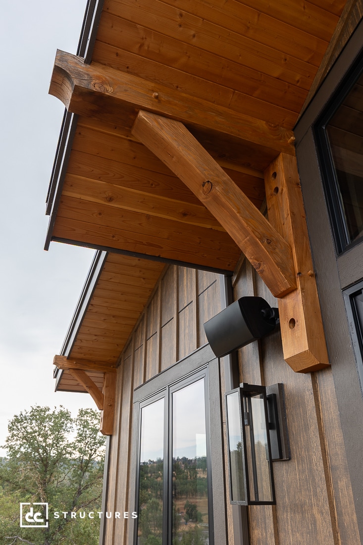 Close-up of a rustic wooden overhang with sturdy beams above large windows on a modern cabin exterior. Trees and sky are visible in the background.