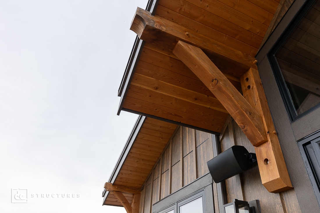 Close-up of a modern wooden awning with exposed beams and metal brackets above a window. The siding features vertical wooden panels. The sky is overcast.