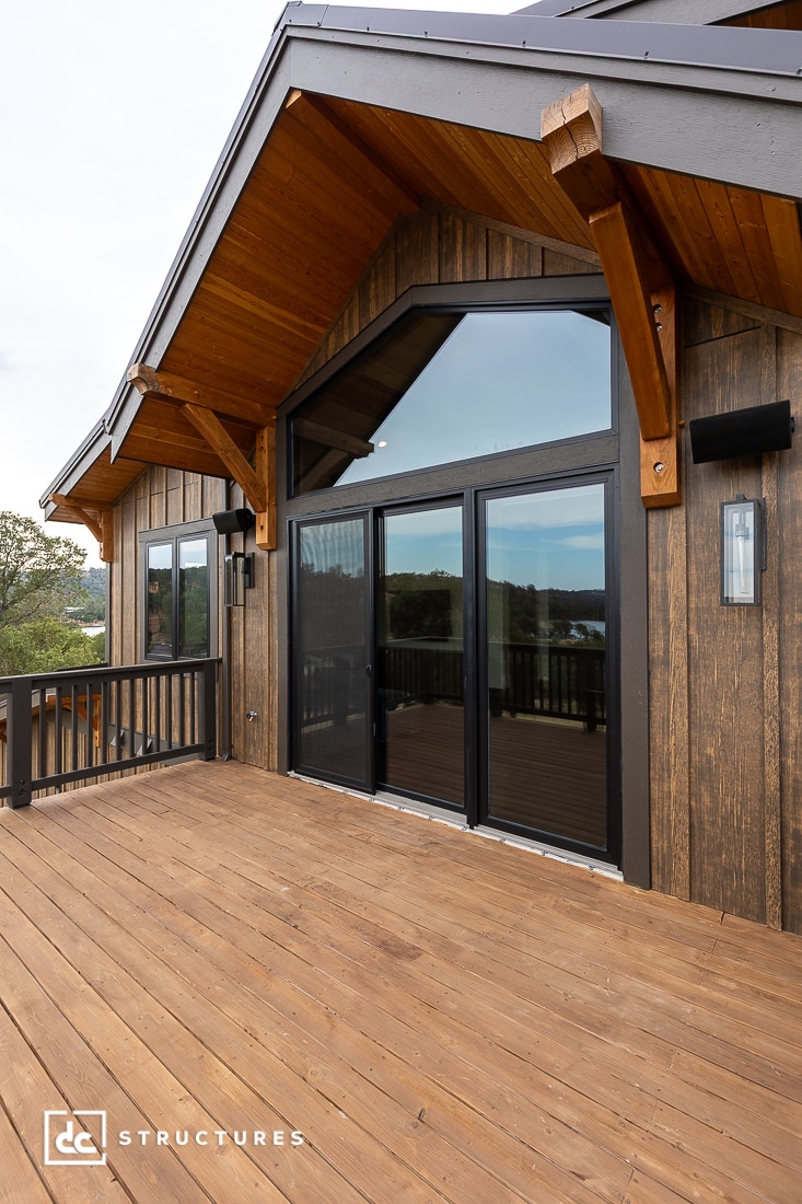 A spacious wooden deck with black-framed glass doors and large windows, featuring exposed timber beams and wood siding. The deck overlooks a scenic outdoor view.
