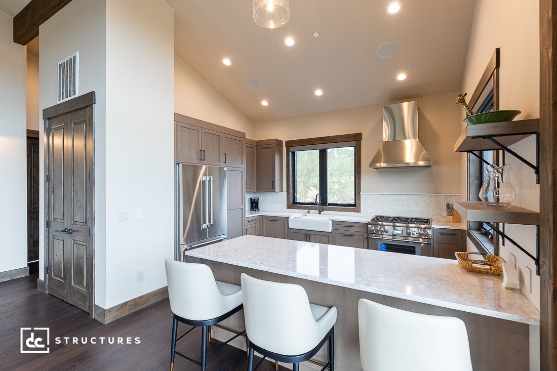 Modern kitchen with gray cabinets, stainless steel appliances, a marble island with three white chairs, large windows, open shelving, wood accents, and recessed lighting.