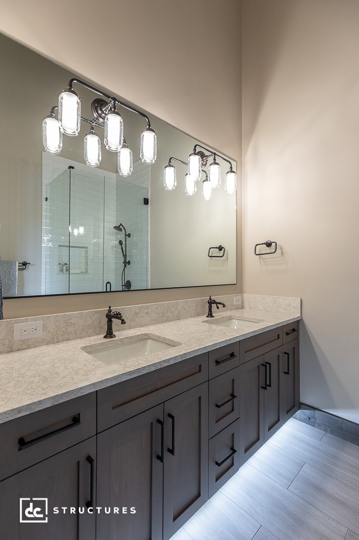 Modern bathroom with a double vanity, marble countertop, two sinks, dark wood cabinets, large wall mirror, and decorative light fixtures above the mirror. A glass-enclosed shower is reflected in the mirror.