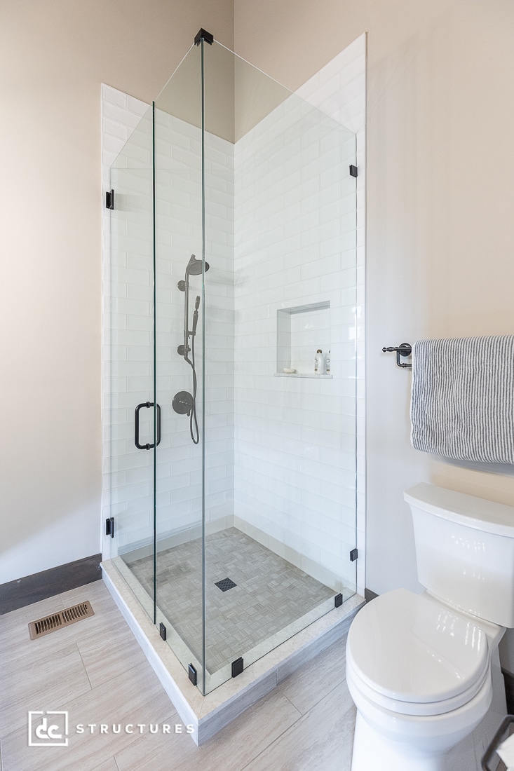 A modern bathroom with a glass-enclosed shower featuring white subway tiles, a rainfall showerhead, built-in shelf, beige floor tiles, a white toilet, and a towel hanging on a nearby rack.