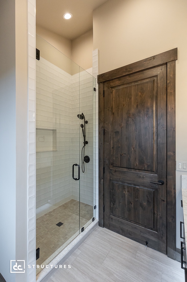 A modern bathroom featuring a glass-enclosed shower with white subway tiles, black shower fixtures, and a rustic dark wooden door next to it. Light-colored floor tiles complete the space.