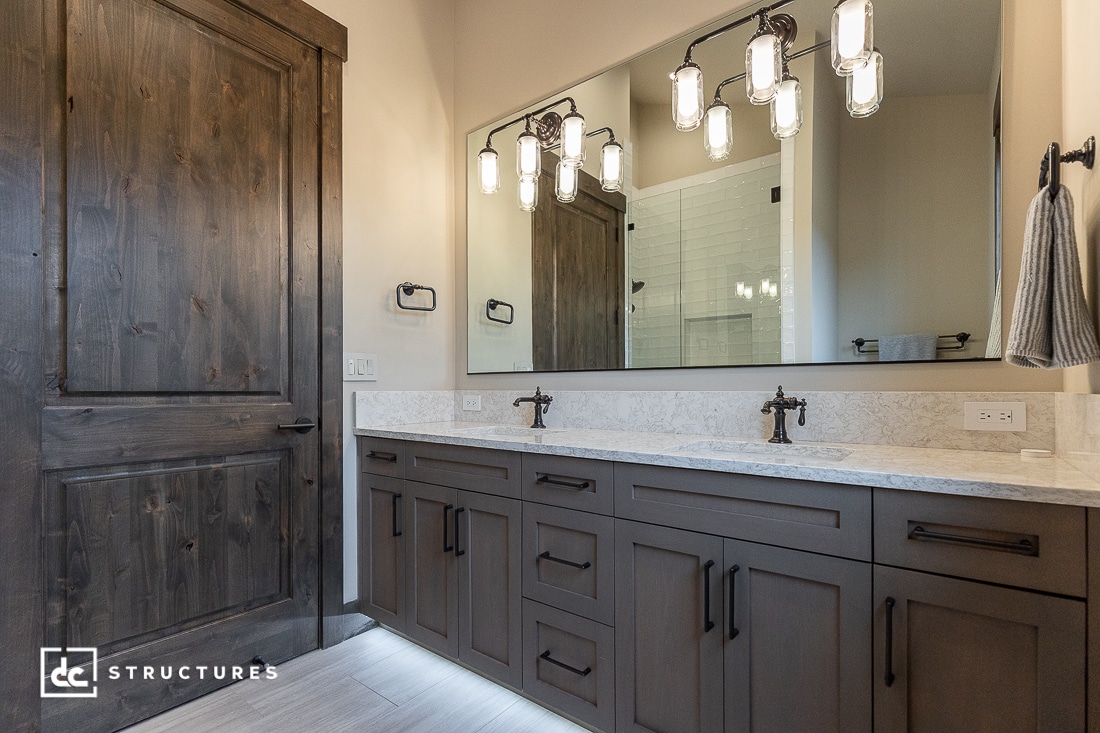 Modern bathroom with double sinks, dark wood cabinets, a large mirror, industrial-style light fixtures, and a wooden door. A towel hangs on the wall, and there's a glimpse of a tiled shower in the reflection.
