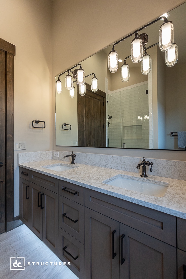 A modern bathroom with a double-sink vanity, marble countertop, dark wood cabinets, a large mirror, and stylish light fixtures above. A glass shower enclosure is visible in the background.