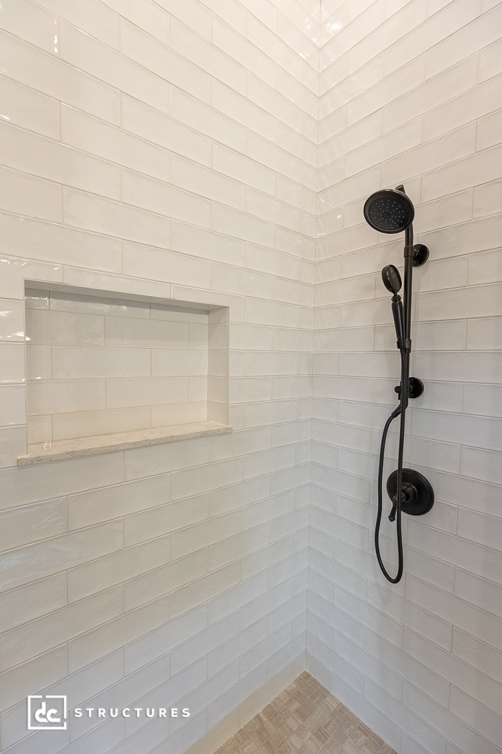 Close-up of a modern shower with glossy white subway tiles, a built-in recessed shelf, and a matte black adjustable showerhead with a handheld attachment. The floor is tiled in light beige tones.