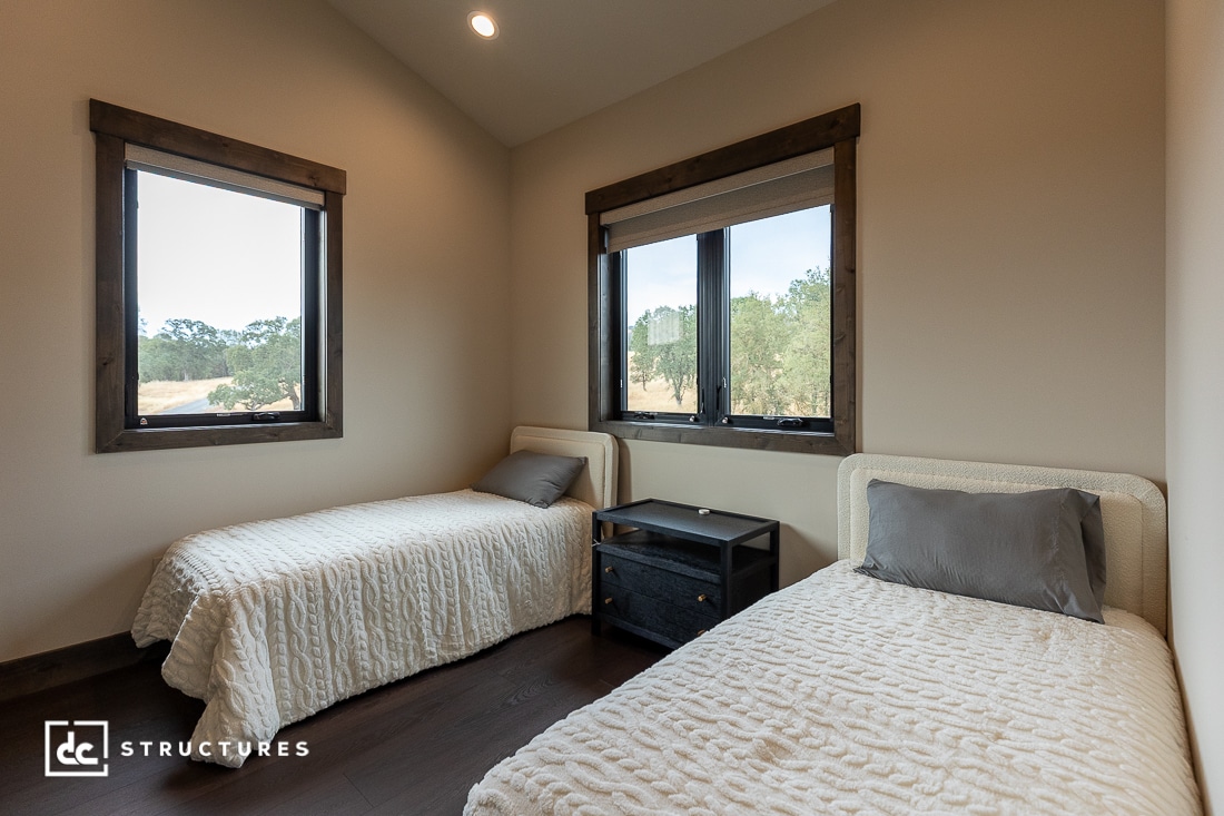 A cozy bedroom with two twin beds, each with a white textured bedspread and gray pillow, separated by a black nightstand, beneath two large windows with a view of trees outside.
