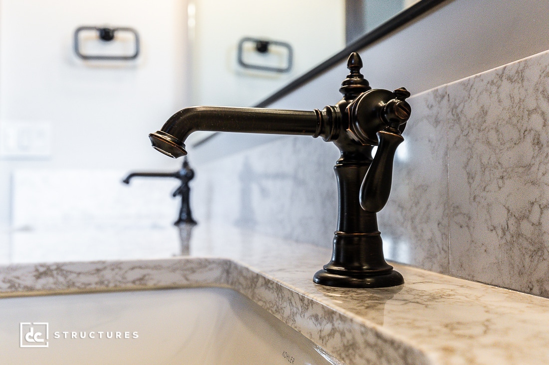 Close-up of an ornate, dark bronze bathroom faucet with a curved spout, mounted on a marble countertop. A matching faucet and reflection are visible in the background.