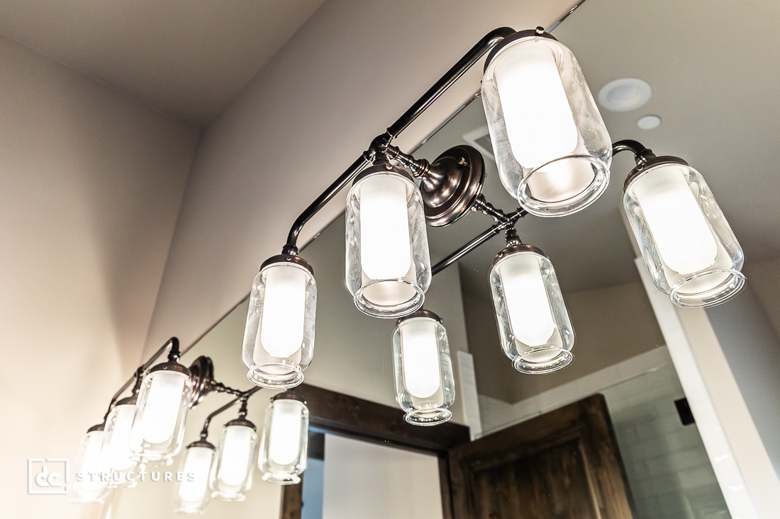 A row of modern, cylindrical glass light fixtures is mounted above a bathroom mirror, reflecting their warm glow. The background includes part of a wooden door and tiled wall.