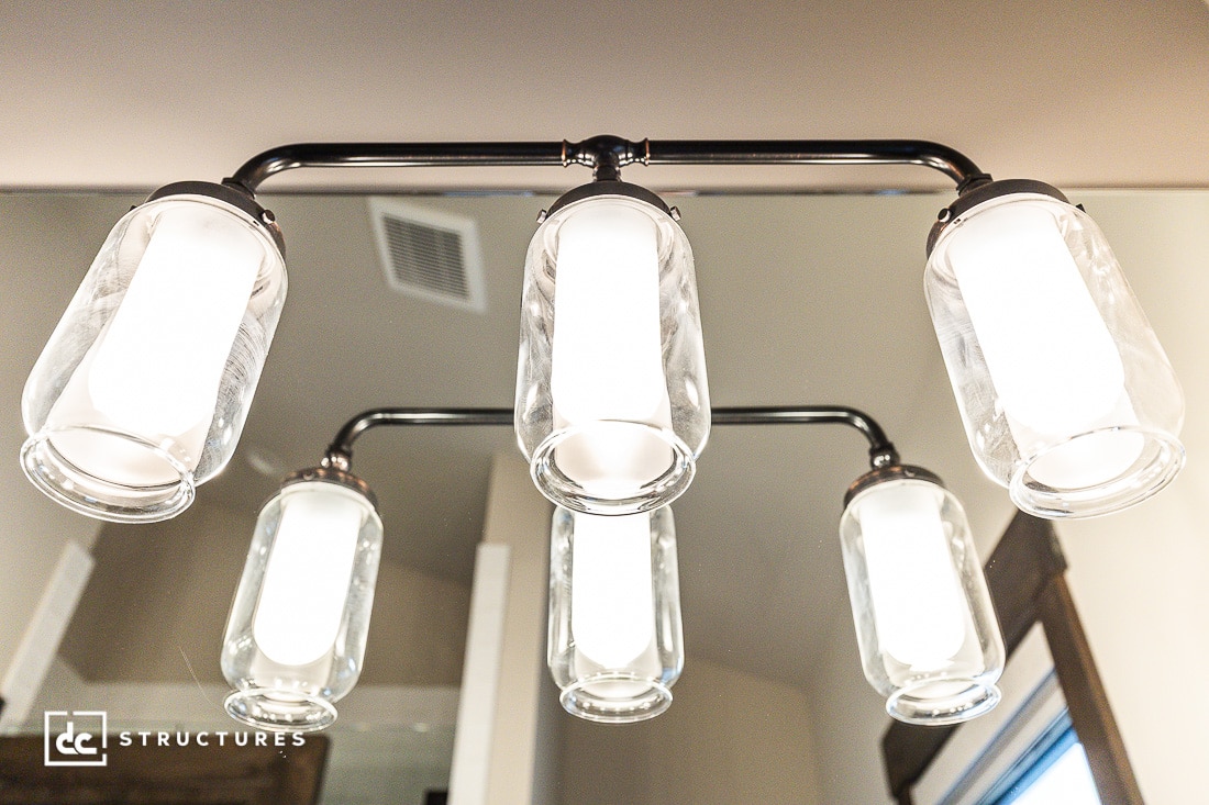 Five mason jar-style light fixtures with bright bulbs are mounted on a chrome bar above a bathroom mirror, reflecting against a beige wall and ceiling vent.
