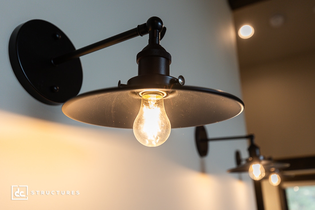 Close-up of a lit wall-mounted industrial-style sconce with a round black metal shade and exposed light bulb, with another similar sconce out of focus in the background.