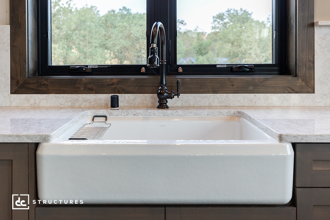 A farmhouse-style white kitchen sink with a black vintage faucet, set in a marble countertop beneath a large window with a wooden frame, overlooking greenery outside.