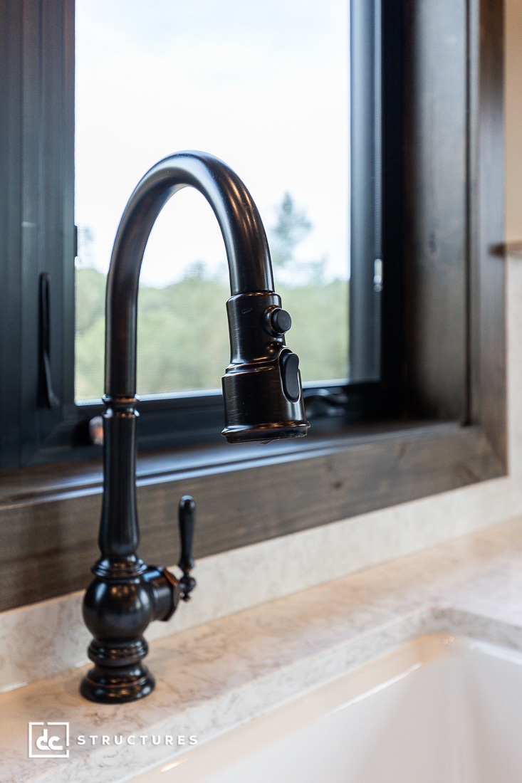 A close-up of a black kitchen faucet with a curved spout and handle, set against a marble countertop. A large window with a dark wood frame is in the background, showing greenery outside.