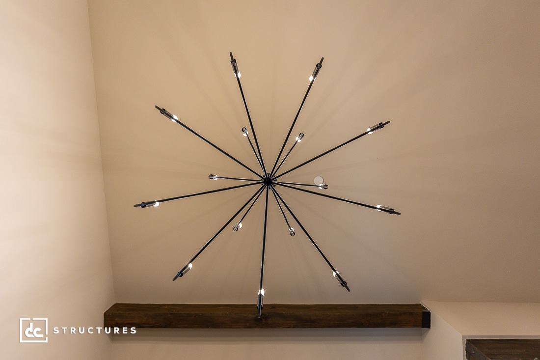 A modern black starburst chandelier with exposed bulbs hangs from a beige ceiling, casting radial shadows. A wooden beam spans the ceiling below.