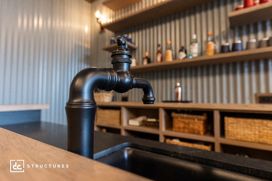 Close-up of a black kitchen faucet over a sink, with shelves holding bottles and baskets in the background. The wall behind features corrugated metal paneling.