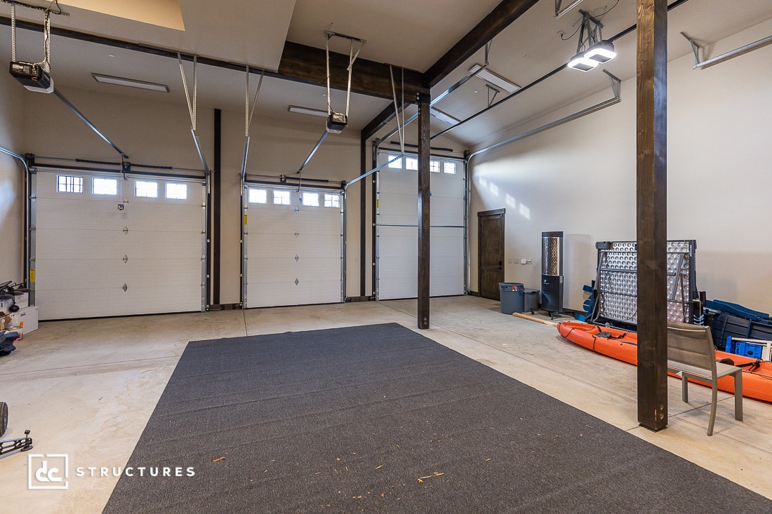 A spacious, well-lit garage with three white overhead doors, a gray carpet in the center, a red kayak, storage shelves, a chair, and various tools and equipment along the right wall.