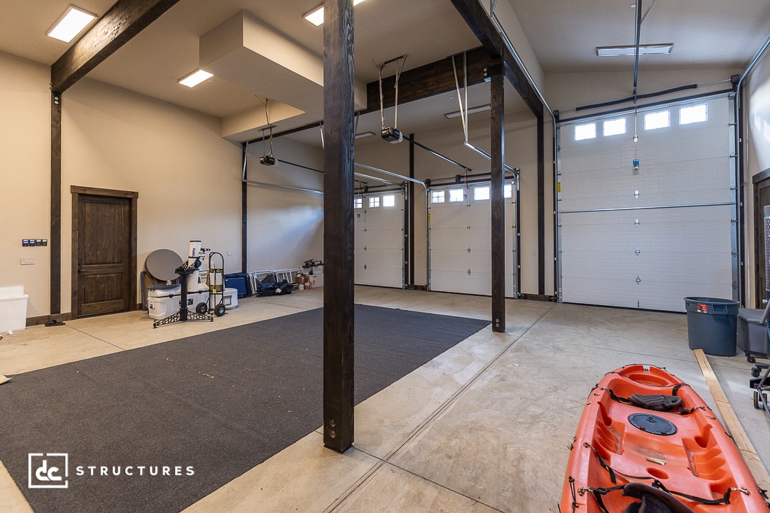 A spacious, modern garage with three large white garage doors, exposed wooden beams, storage bins, a bright orange kayak, and various equipment against the walls. The floor is concrete with a dark mat in the center.