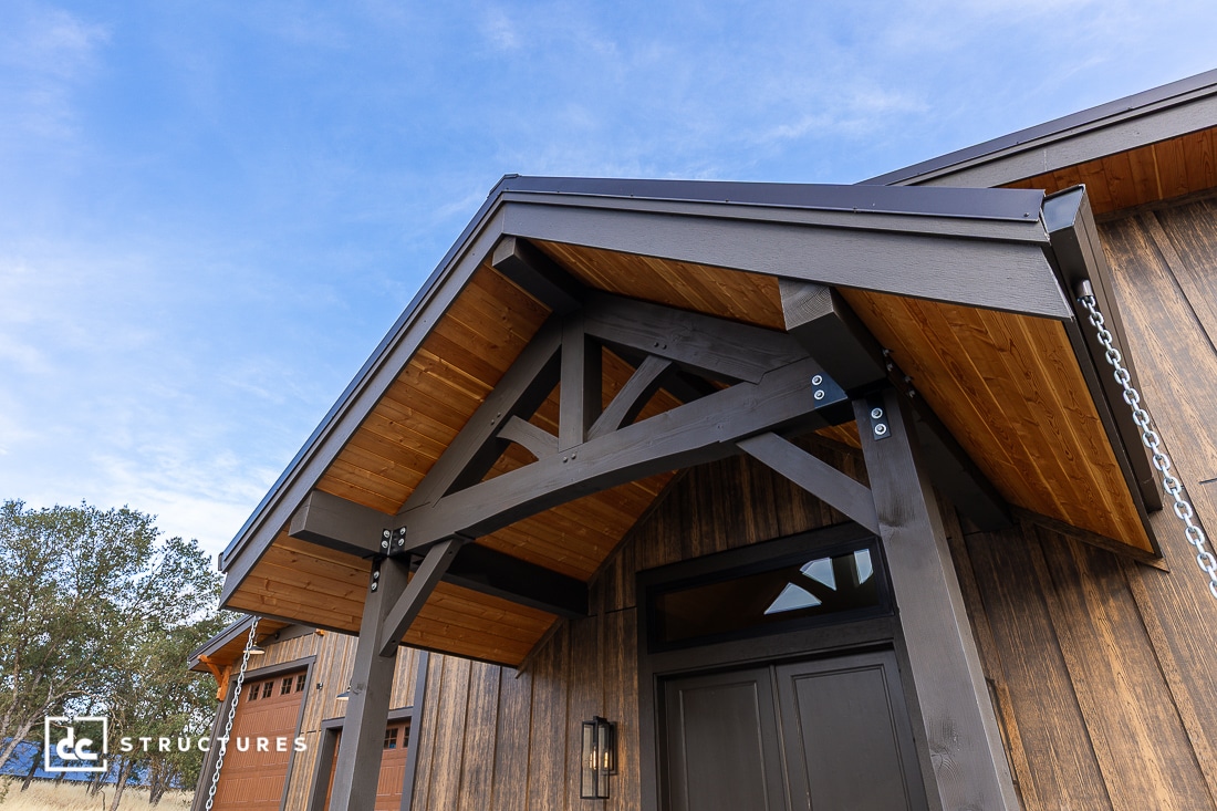 A close-up view of a modern wooden house entrance with a covered gabled porch, dark beams, and natural wood paneling under a blue sky.